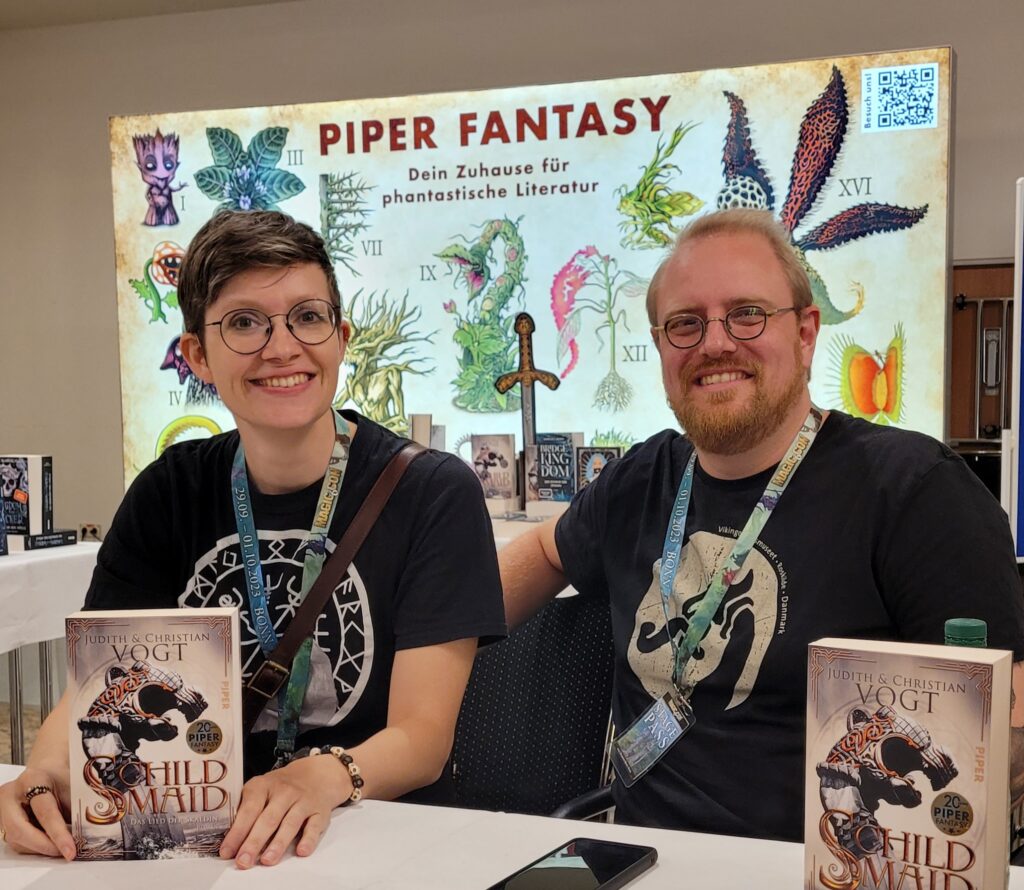 Judith C. Vogt und Christian Vogt sitzen mit Ausgaben ihres Buches "Schildmaid"" vor einem Poster von Piper Fantasy - "Dein Zuhause für phantastische Literatur. 